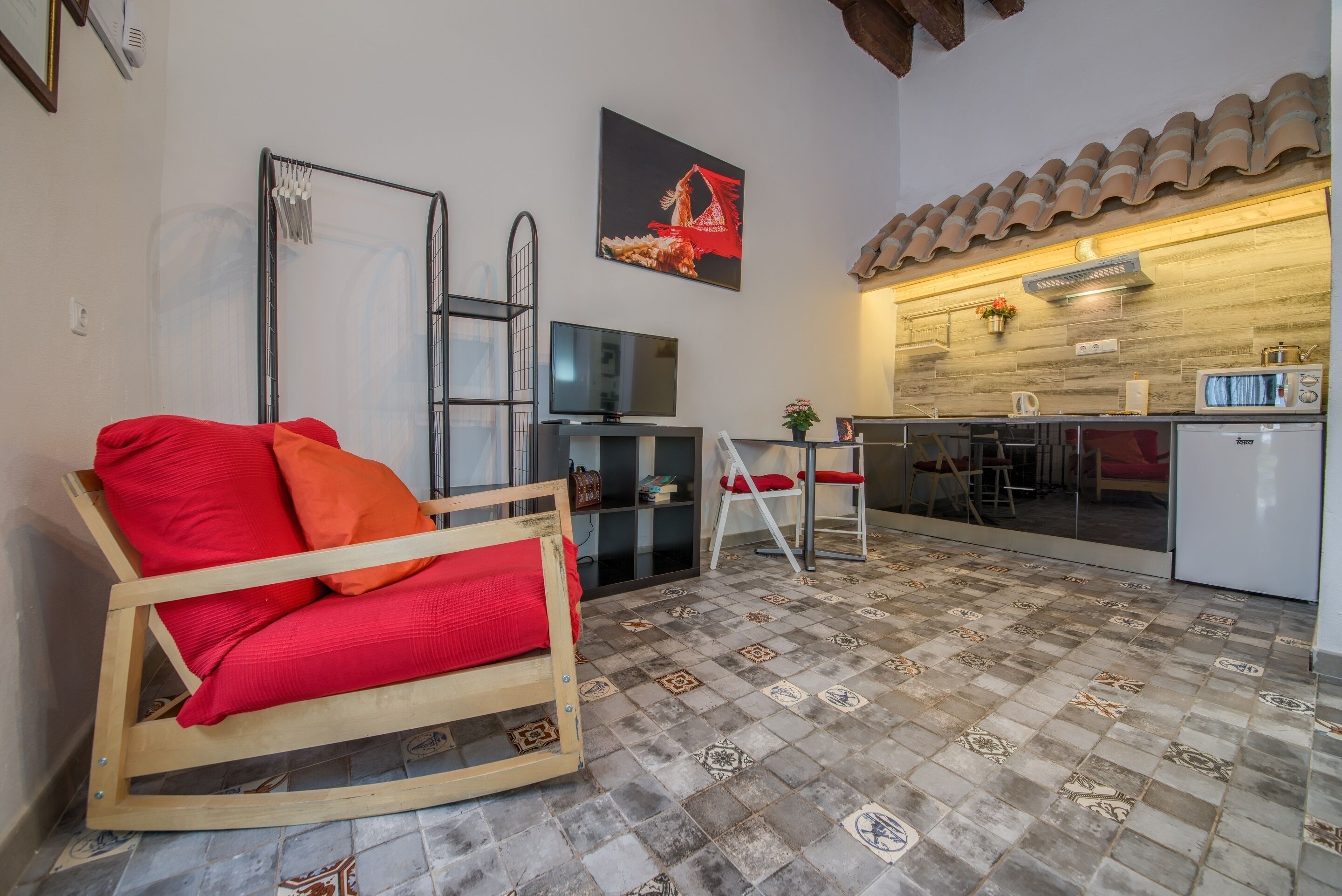 A compact living space with a wooden chair, a TV, a kitchen area with a microwave, and a tiled floor.