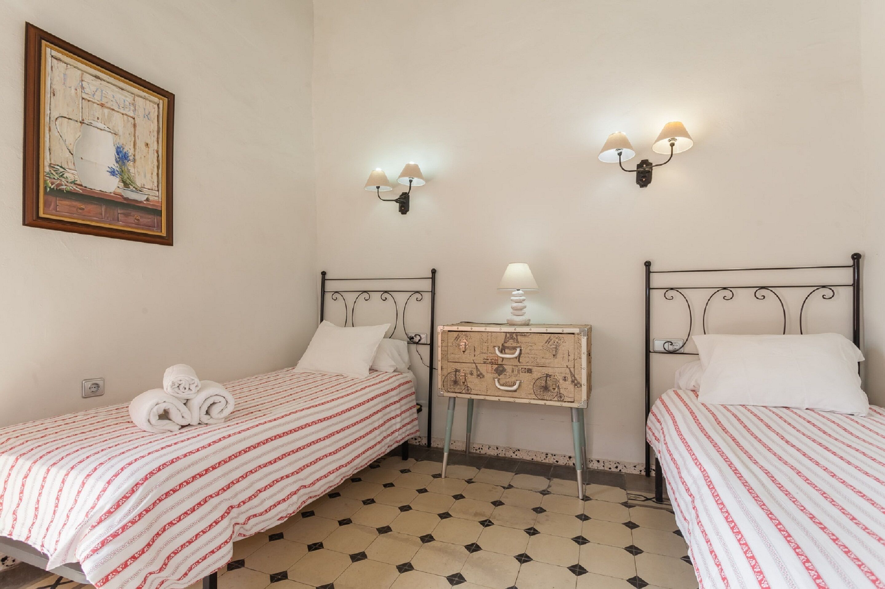 A room with two beds, each with a striped bedspread, a small bedside table with a lamp, and wall-mounted light fixtures.