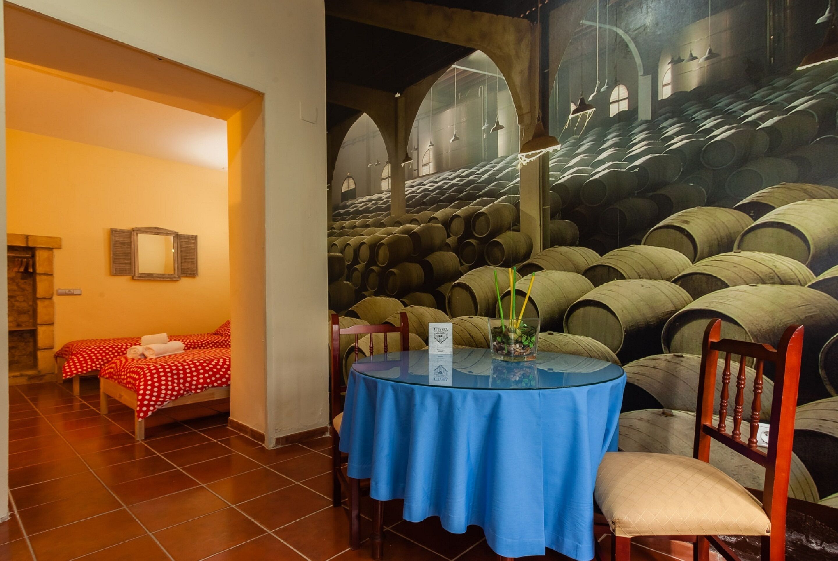 Una habitación con una cama, una mesa con mantel azul y un mural en la pared de barriles de vino.