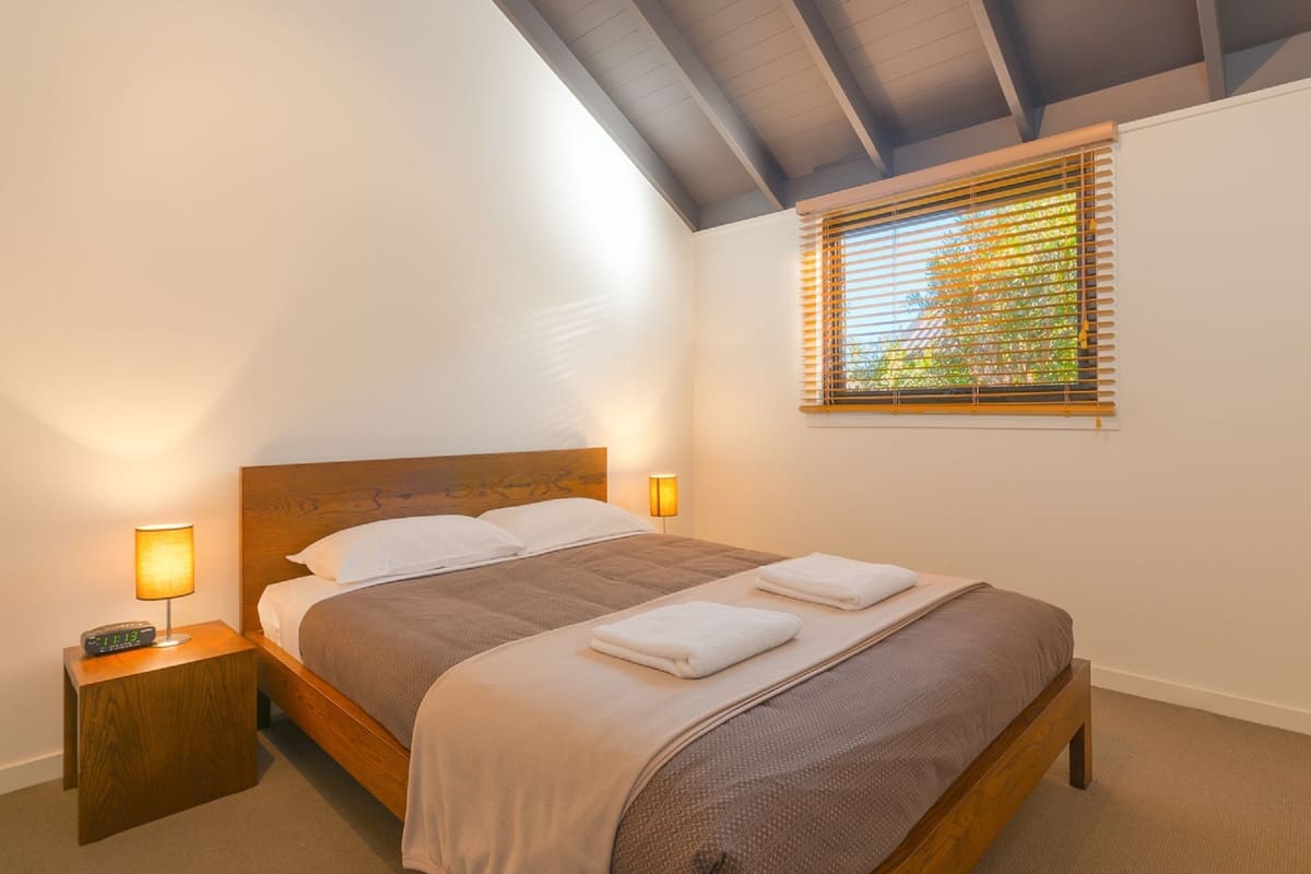 Un dormitorio con una cama de madera, dos mesitas de noche con lámparas, una ventana con persianas y un techo inclinado.