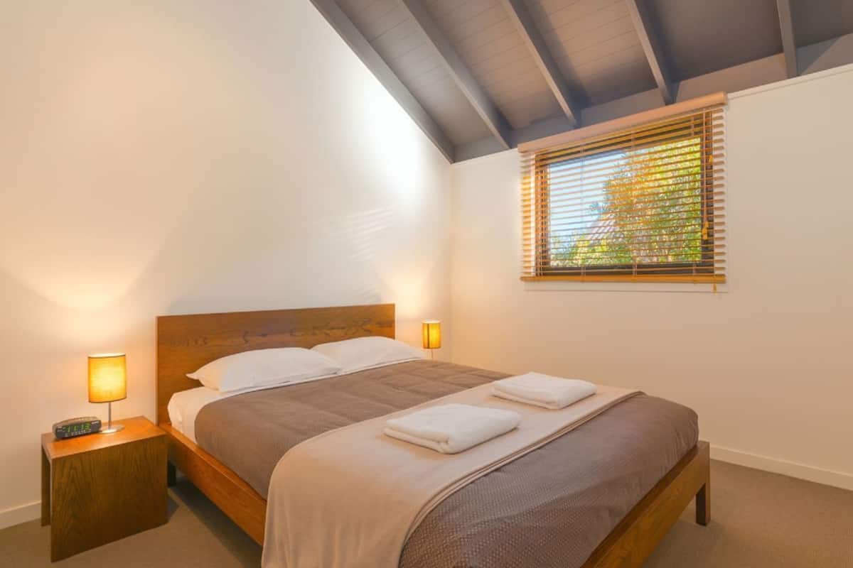 Una cama individual con cabecera de madera, una mesita de noche con lámpara, una ventana con persianas y un estante fijado a la pared.