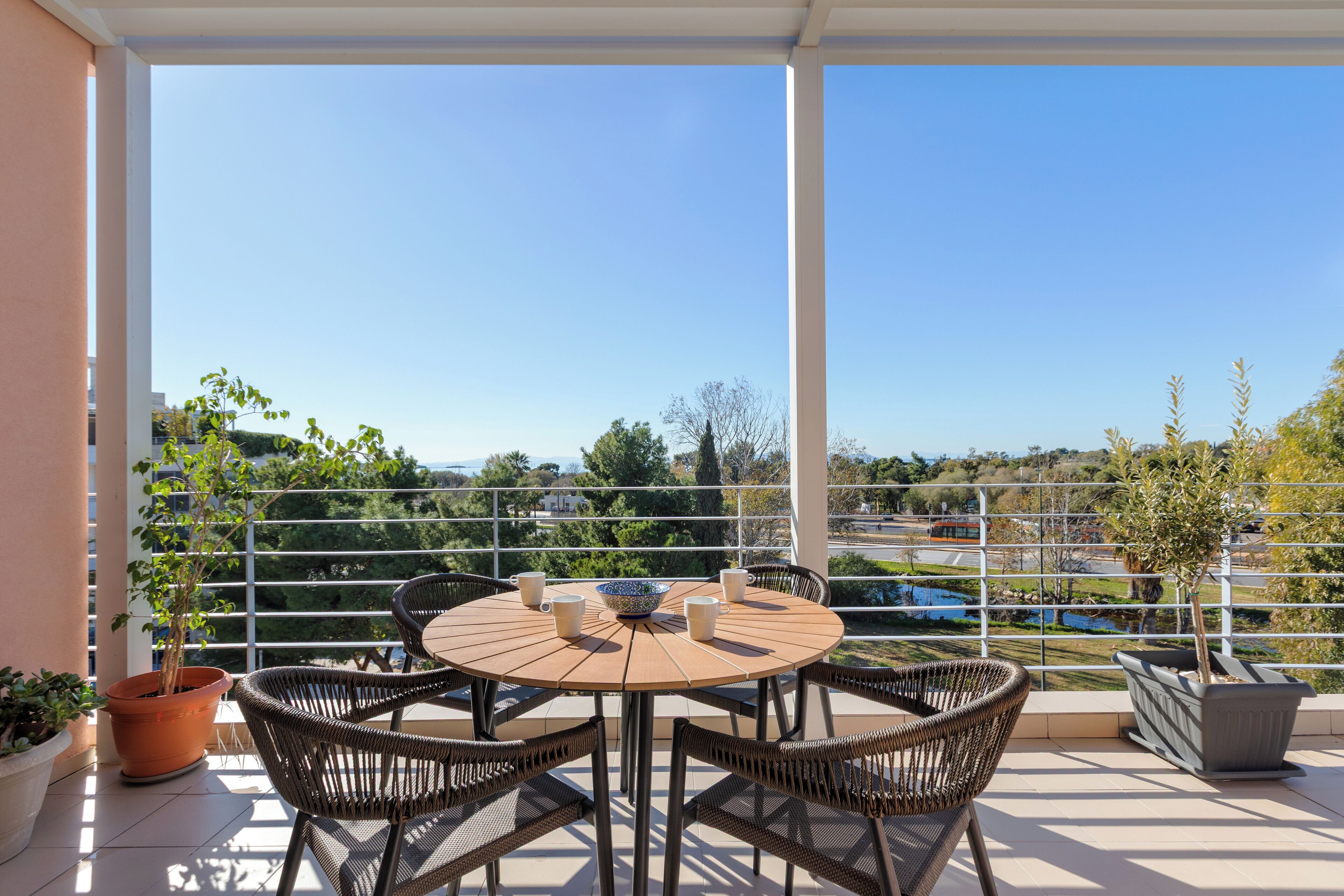 A round table with four wicker chairs, a potted plant, and two coffee cups on a balcony with a view of a lake and buildings.