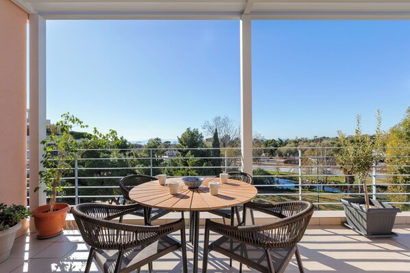 A round table with four wicker chairs, a potted plant, and two coffee cups on a balcony with a view of a lake and buildings. - Minavra Hotel (Vari-Voula-Vouliagmeni)