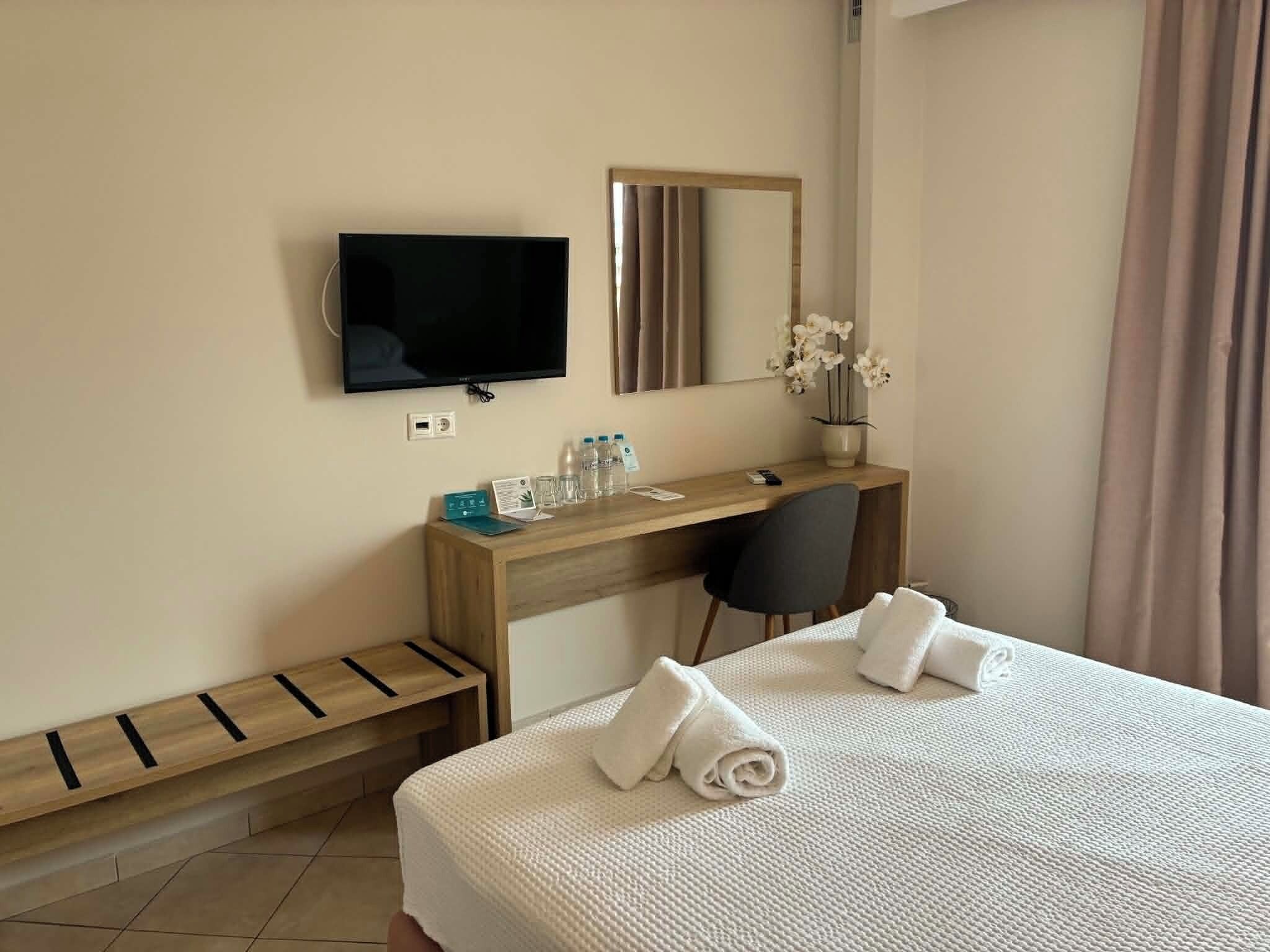 Een hotelkamer met een bed, een bureau, een televisie, een spiegel, een stoel en een klein bankje.