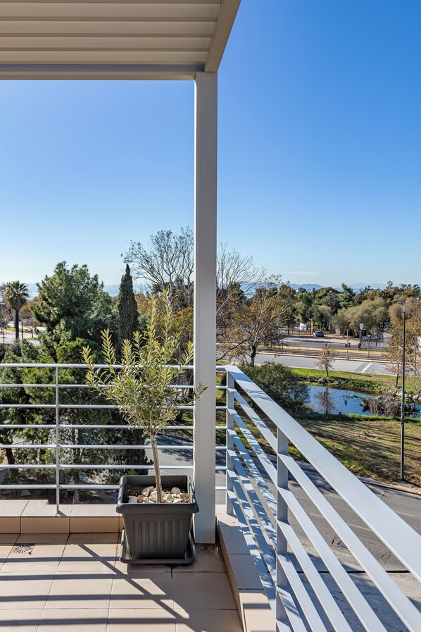 A balcony with a view of a park, a road, and distant mountains. - Minavra Hotel (Vari-Voula-Vouliagmeni)