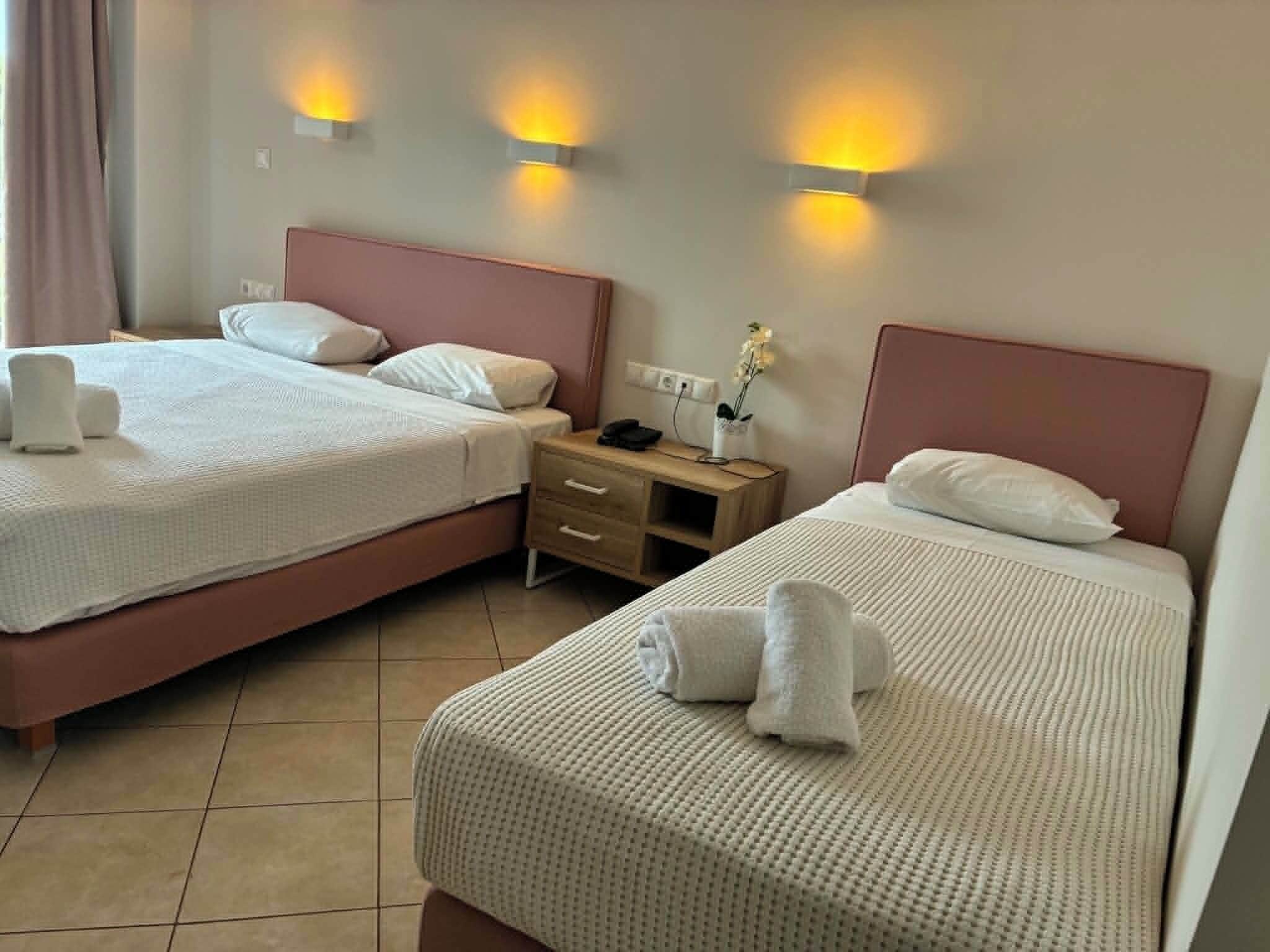 Een hotelkamer met twee bedden, elk voorzien van witte beddengoed en kussens, een houten nachtkastje met een telefoon en een vaas met bloemen, plus wandlampen.
