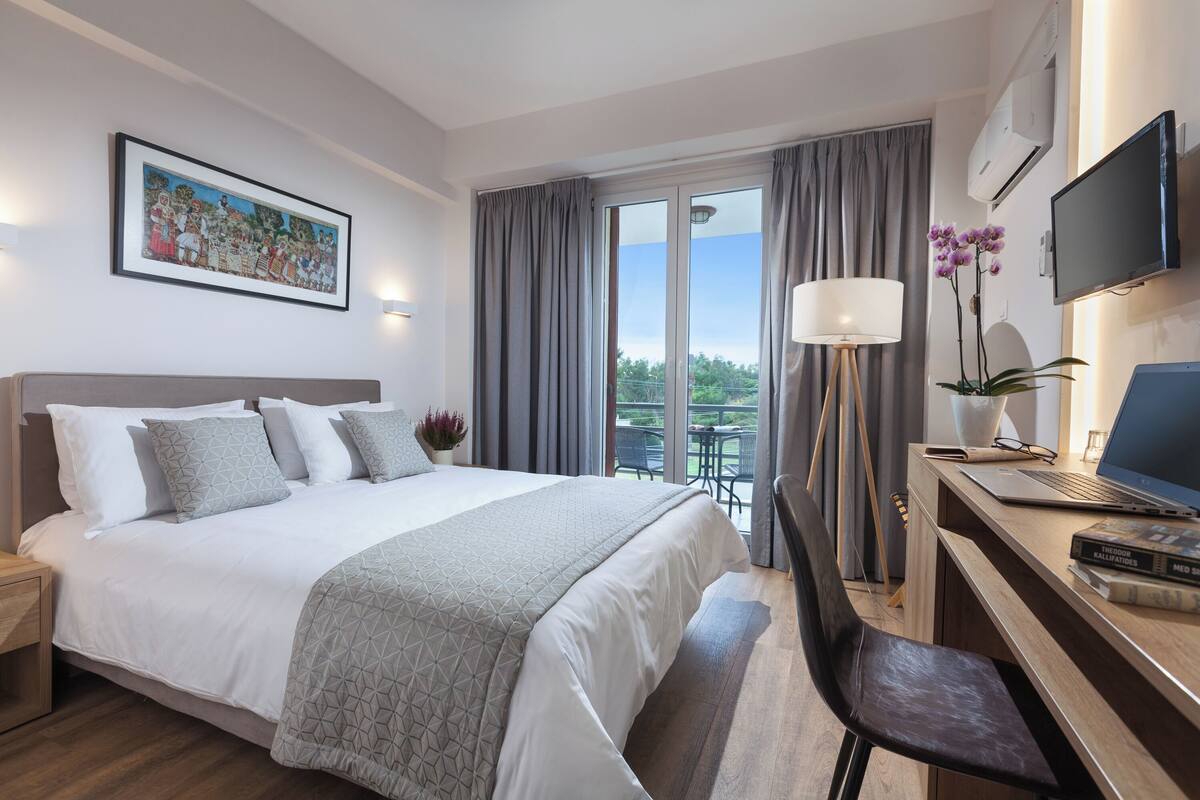 A hotel room with a bed, a desk with a laptop, a TV, and a balcony with curtains.