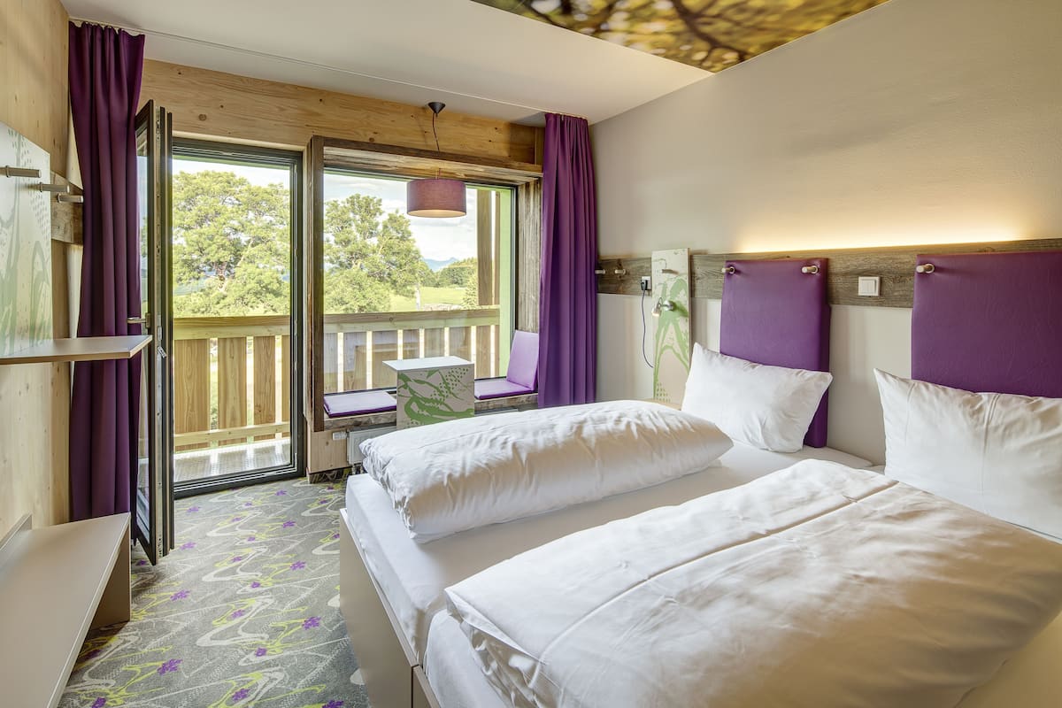 Habitación de hotel con dos camas, techo de madera, balcón con cortinas moradas y vistas a árboles.