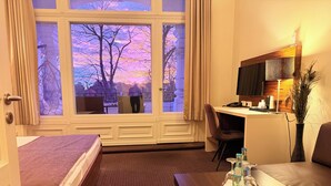 A hotel room with a bed, a desk, a chair, a TV, and a window with a view of trees. - Hotel Excellent (Lübeck)