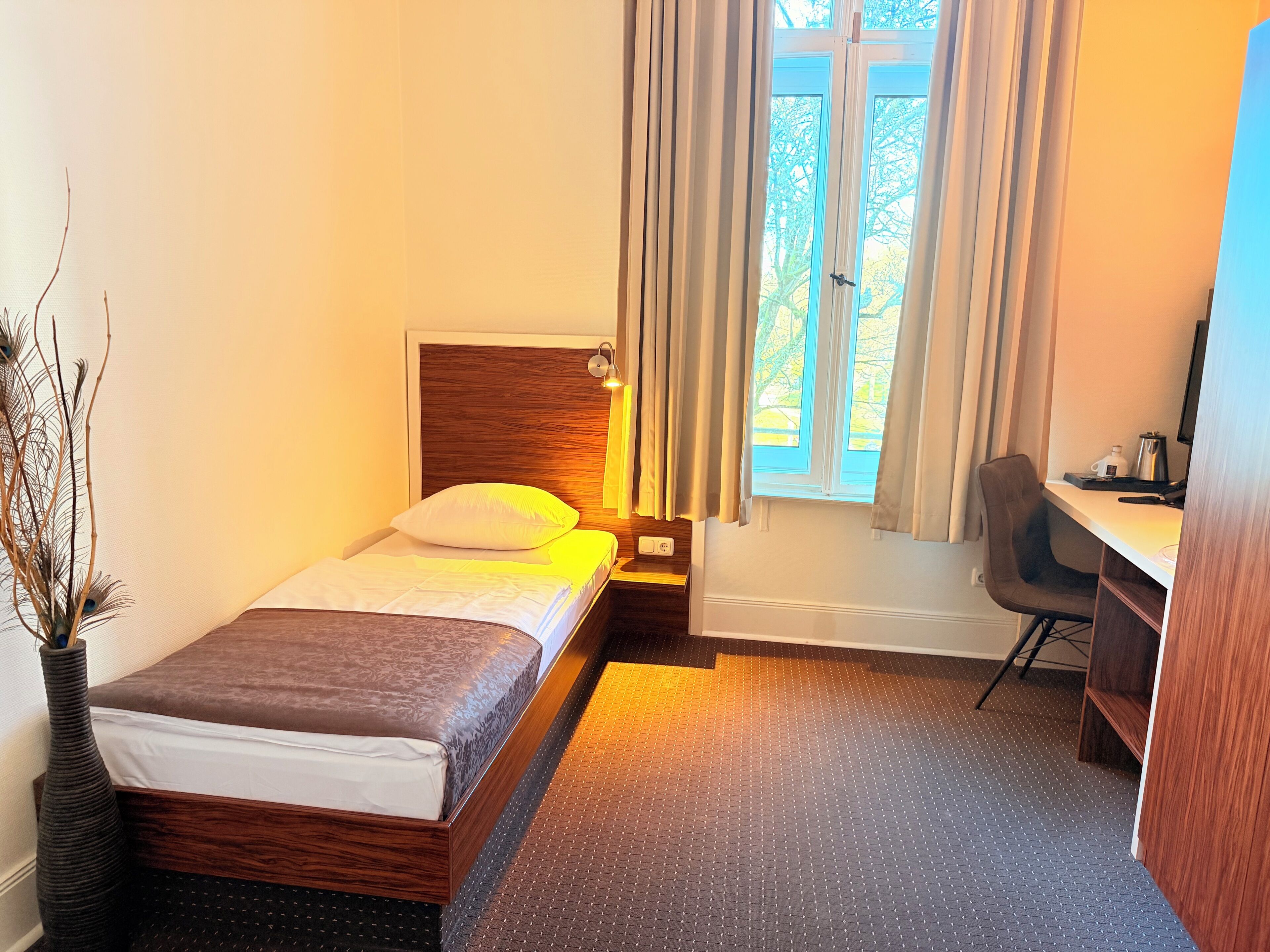 A hotel room with a bed, a desk, a chair, a window with curtains, and a vase with branches.