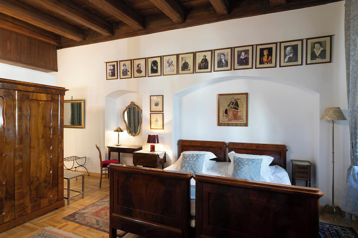 A bedroom with a wooden bed, a desk with a lamp, a chair, and a wardrobe. There are framed pictures on the wall and a painting above the bed.