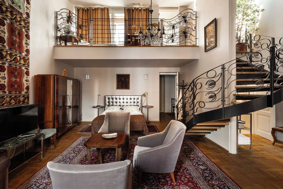 A spacious living room with a high ceiling, a large bed, a staircase with ornate railings, and a television.