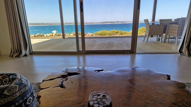 A room with a large glass door leading to a beach view, a wooden coffee table with a glass of drink, and wicker furniture.