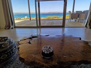 A room with a large glass door leading to a beach view, a wooden coffee table with a glass of drink, and wicker furniture. - Koopalanda Dreaming (Vivonne Bay)