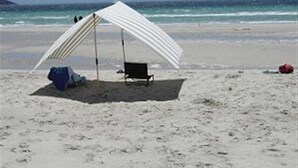 Una playa con una sombrilla blanca que da sombra, una silla de playa negra y un enfriador azul sobre la arena.