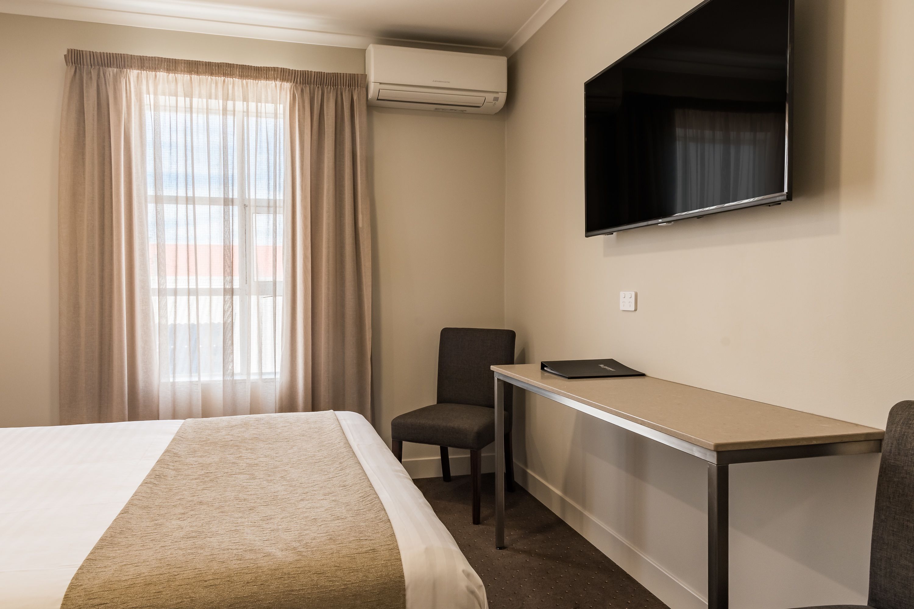 Une chambre d’hôtel équipée d’un lit, d’un bureau, d’une chaise, d’un climatiseur et d’un téléviseur fixé au mur.