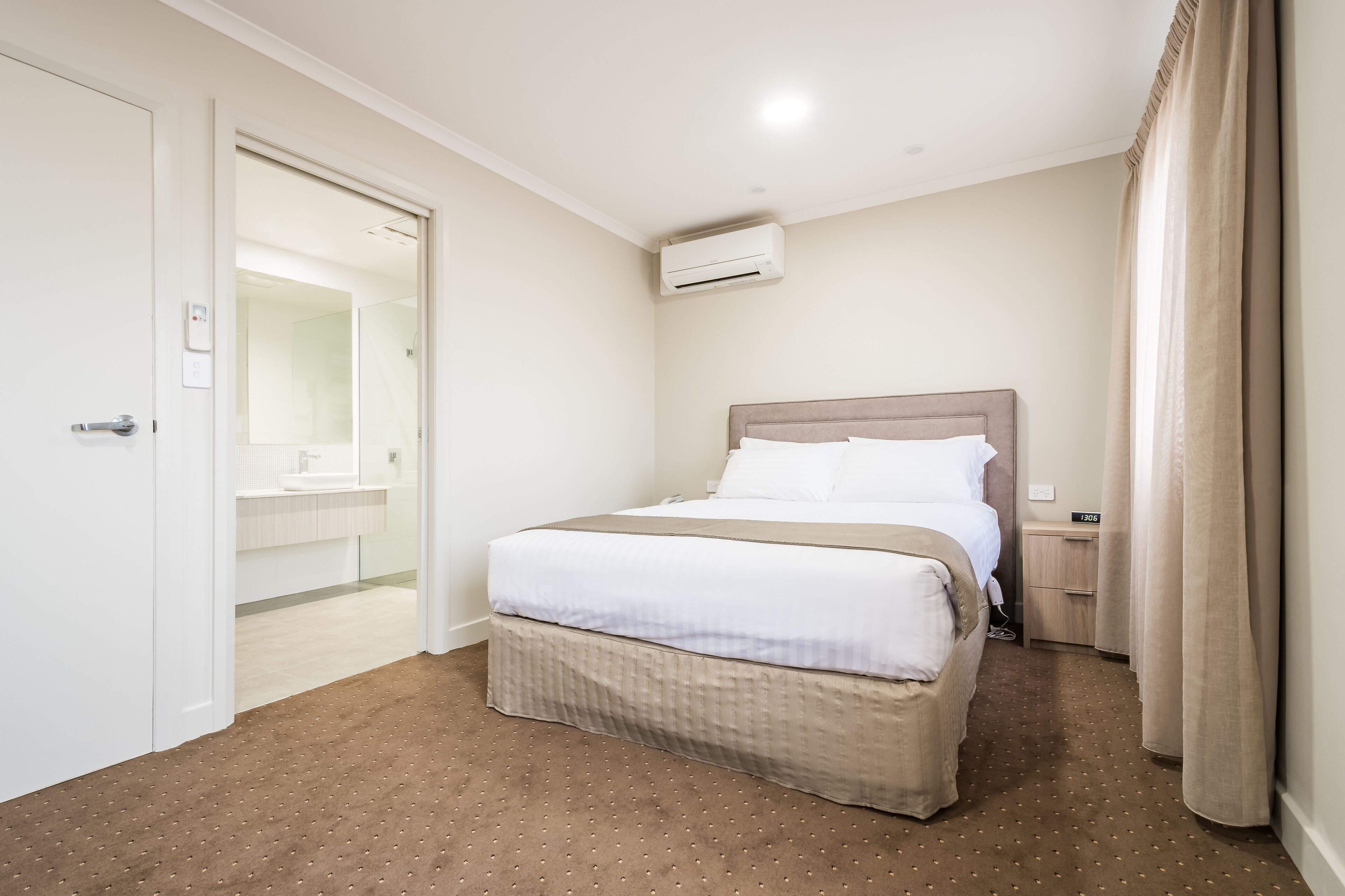 Une chambre d’hôtel avec un lit, une table de chevet, un appareil de climatisation, une salle de bain visible à travers une porte ouverte et un sol recouvert de moquette.