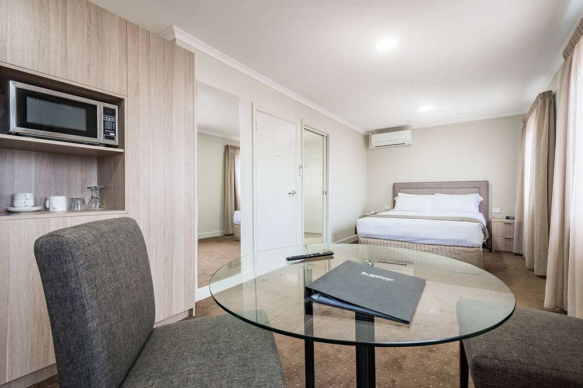 Habitación de hotel con una mesa de vidrio, dos sillones grises, una cama con sábanas blancas, un microondas sobre una estantería de madera y un escritorio pequeño con un control remoto.