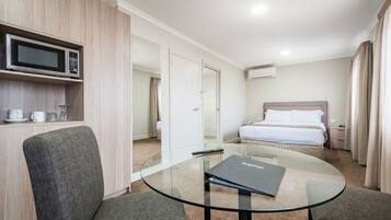 Hotelkamer met glazen tafel, twee grijze fauteuils, bed met witte lakens, magnetron op een houten plank en een klein bureau met afstandsbediening.