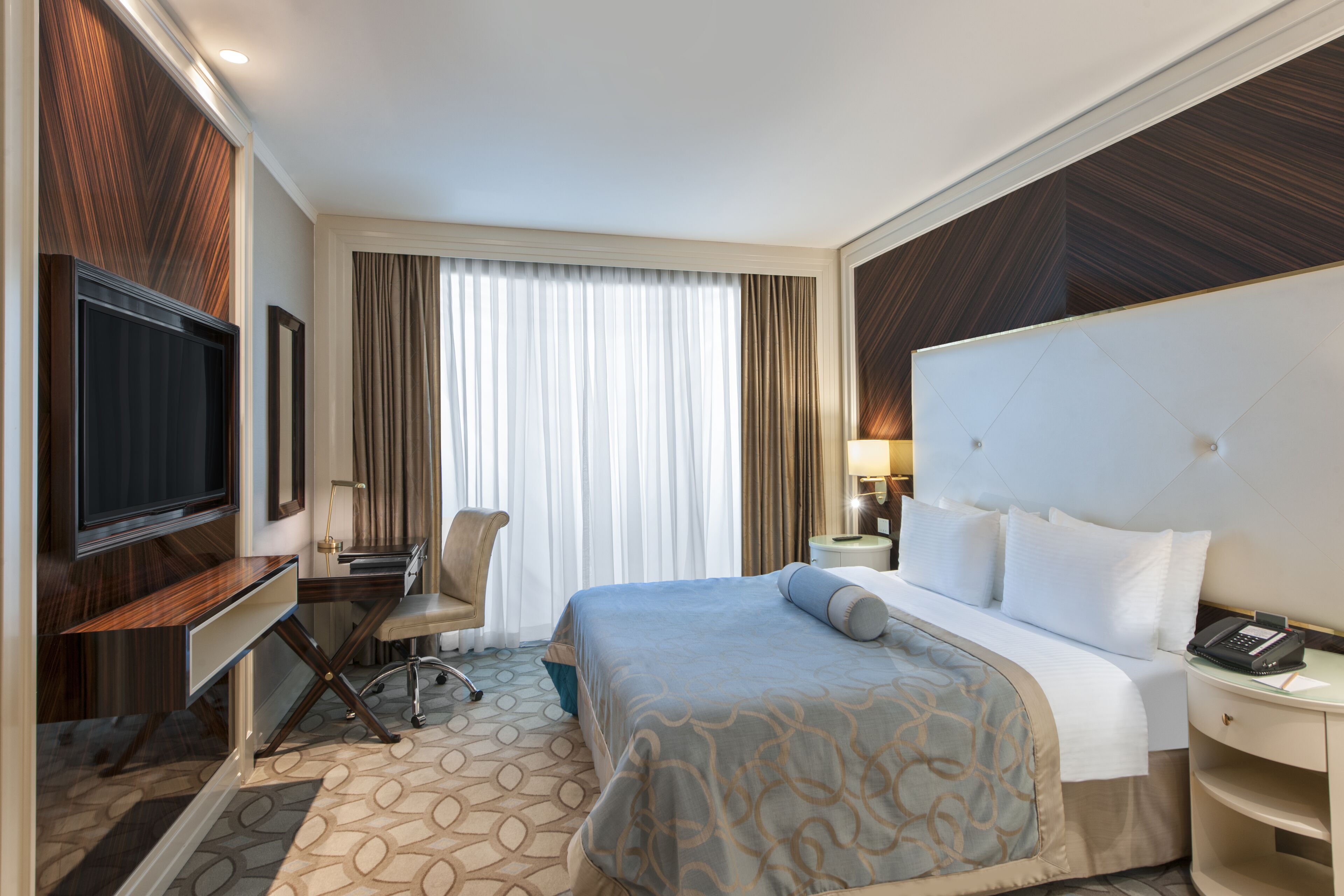 A hotel room with a large bed, a desk with a flat-screen TV, a chair, and a window with curtains.