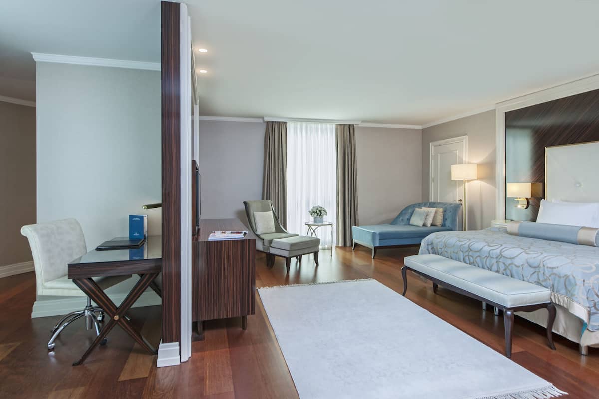 A hotel room with a desk, a chair, a sofa, a bed, and a small table with a lamp.