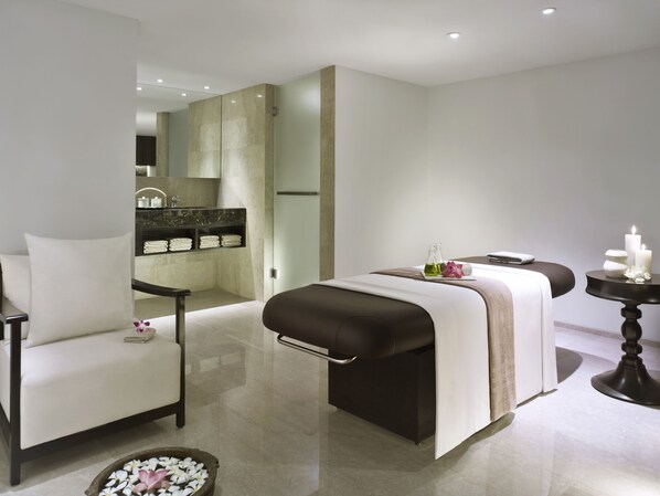 A modern spa room with a massage table, a chair, a small table with candles, and a vanity with a mirror and shelves.