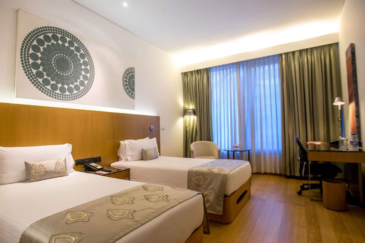 A hotel room with two beds, a desk, a chair, a lamp, and a large window with curtains.