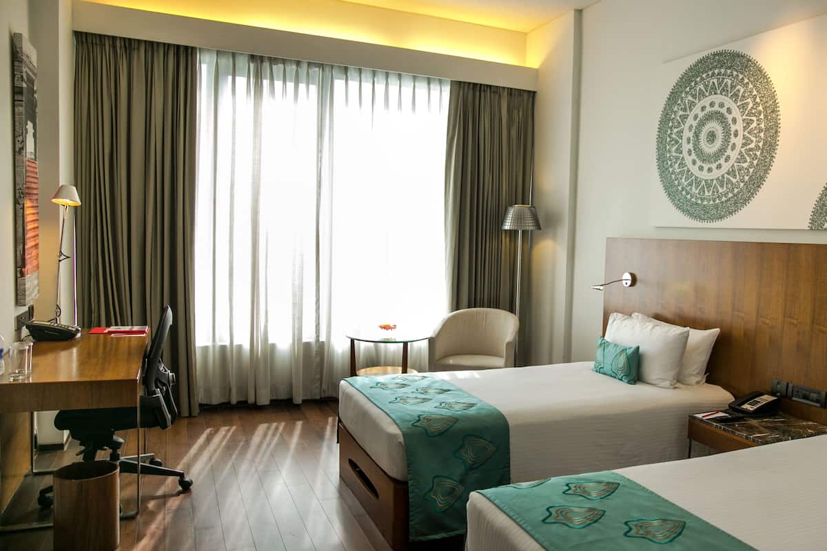 A hotel room with two beds, a desk with a chair, a lamp, and a view of the outdoors through large windows.