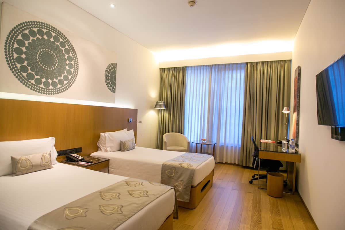 A hotel room with two beds, a desk with a chair, a television, and a large window with curtains.