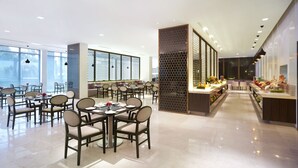 Uma zona de refeições moderna com mesas e cadeiras redondas, um balcão de buffet com comida e vista para a cidade através de grandes janelas.