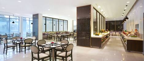 A modern dining area with round tables and chairs, a buffet counter with food, and a view of the city through large windows.