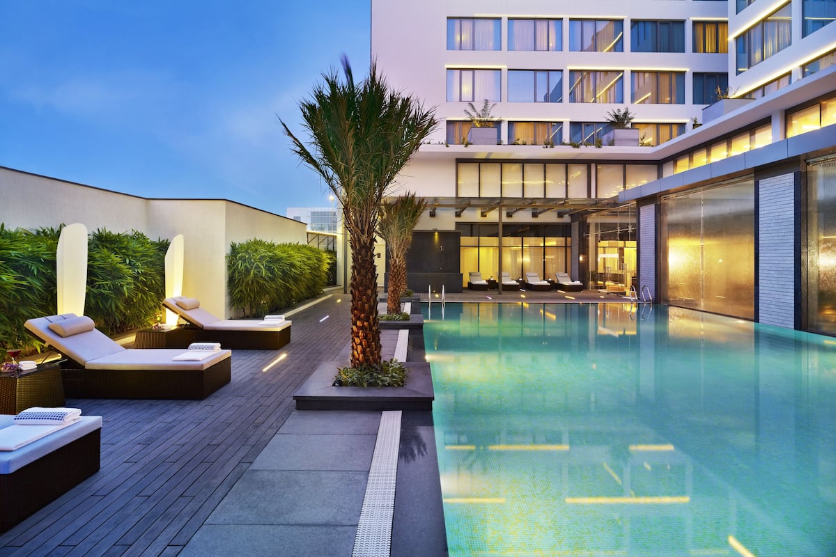 A modern hotel pool area with lounge chairs, a palm tree, and a building with large windows in the background.