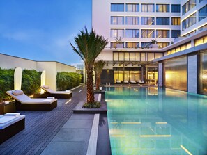 Une piscine moderne d’hôtel, avec des chaises longues, un palmier et, en arrière-plan, un bâtiment doté de grandes fenêtres.