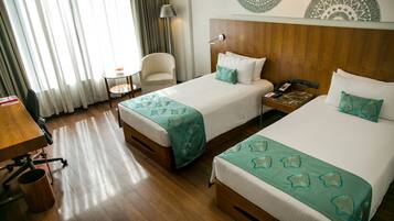 A hotel room with two beds, a wooden desk, a chair, a small table, and a lamp.
