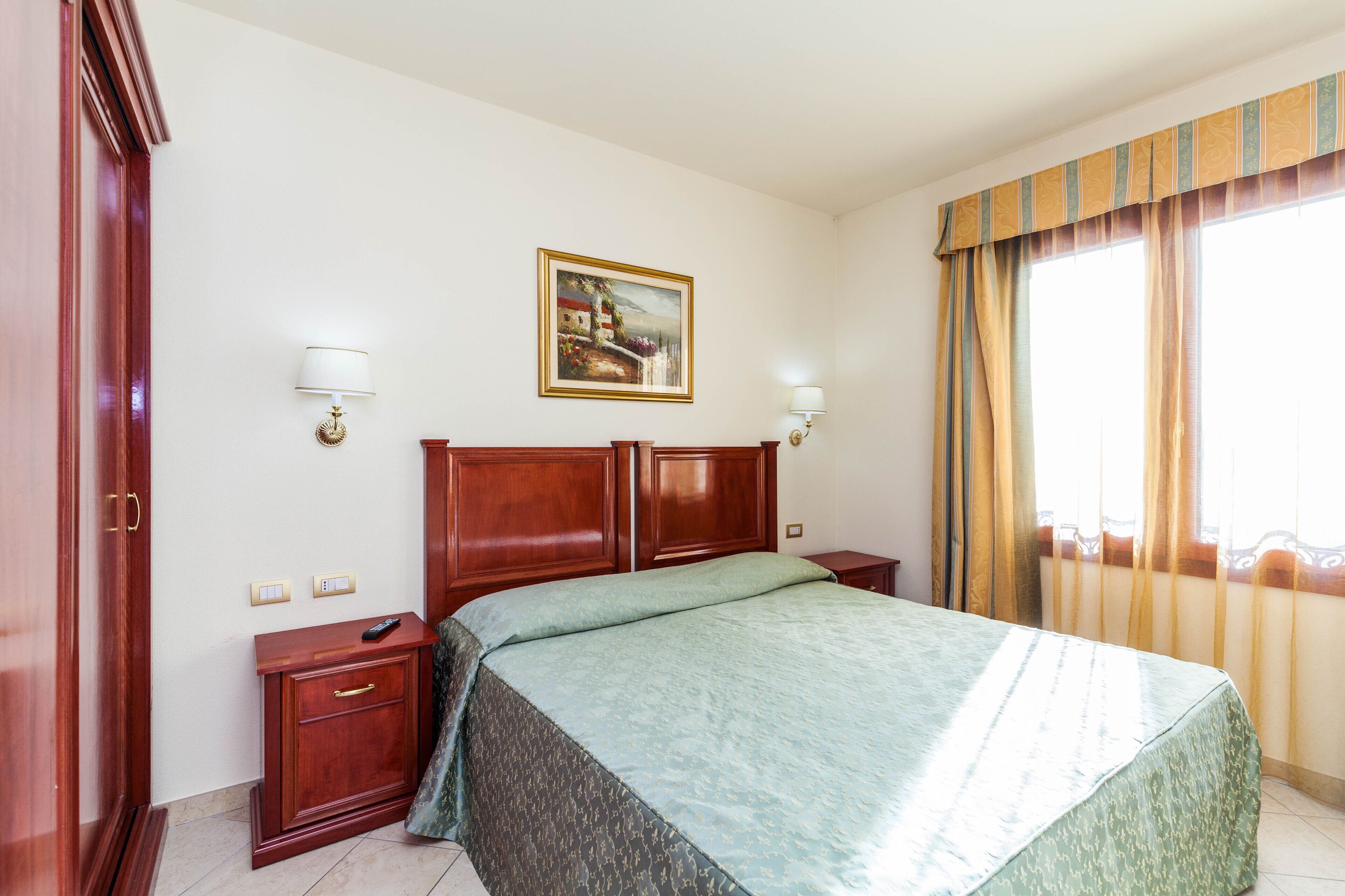 A hotel room with a large bed, wooden headboard, two bedside tables with lamps, a painting on the wall, and a window with curtains.