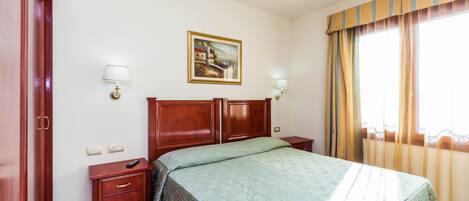 A hotel room with a large bed, wooden headboard, two bedside tables with lamps, a painting on the wall, and a window with curtains.