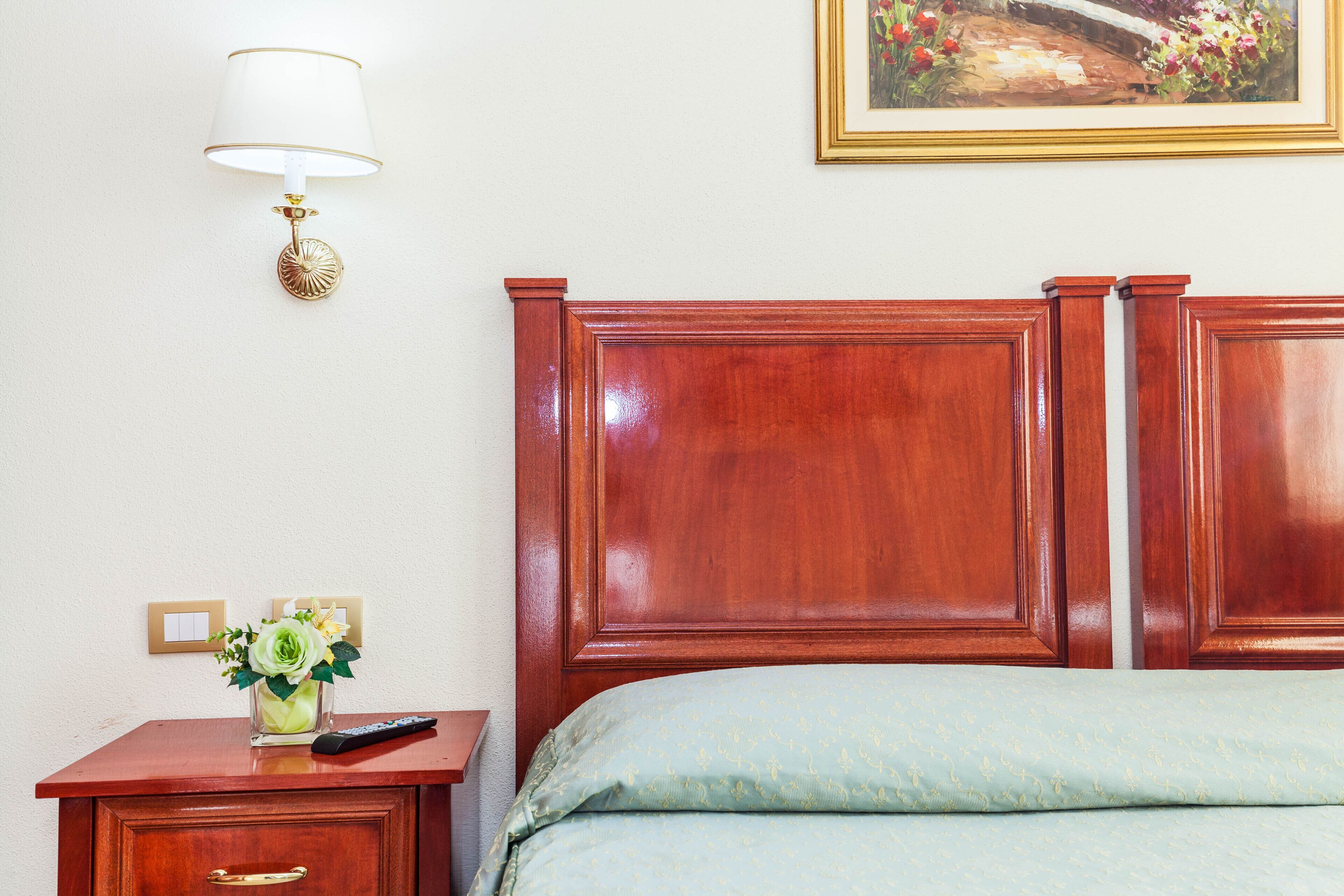 Habitación de hotel con cabecera de madera, mesita de noche con control remoto, un jarrón con flores e imagen enmarcada en la pared.