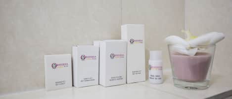 Un baño con pared de mármol, cuatro cajas etiquetadas RHIDEEA, una botella de loción corporal y un vaso con una flor.