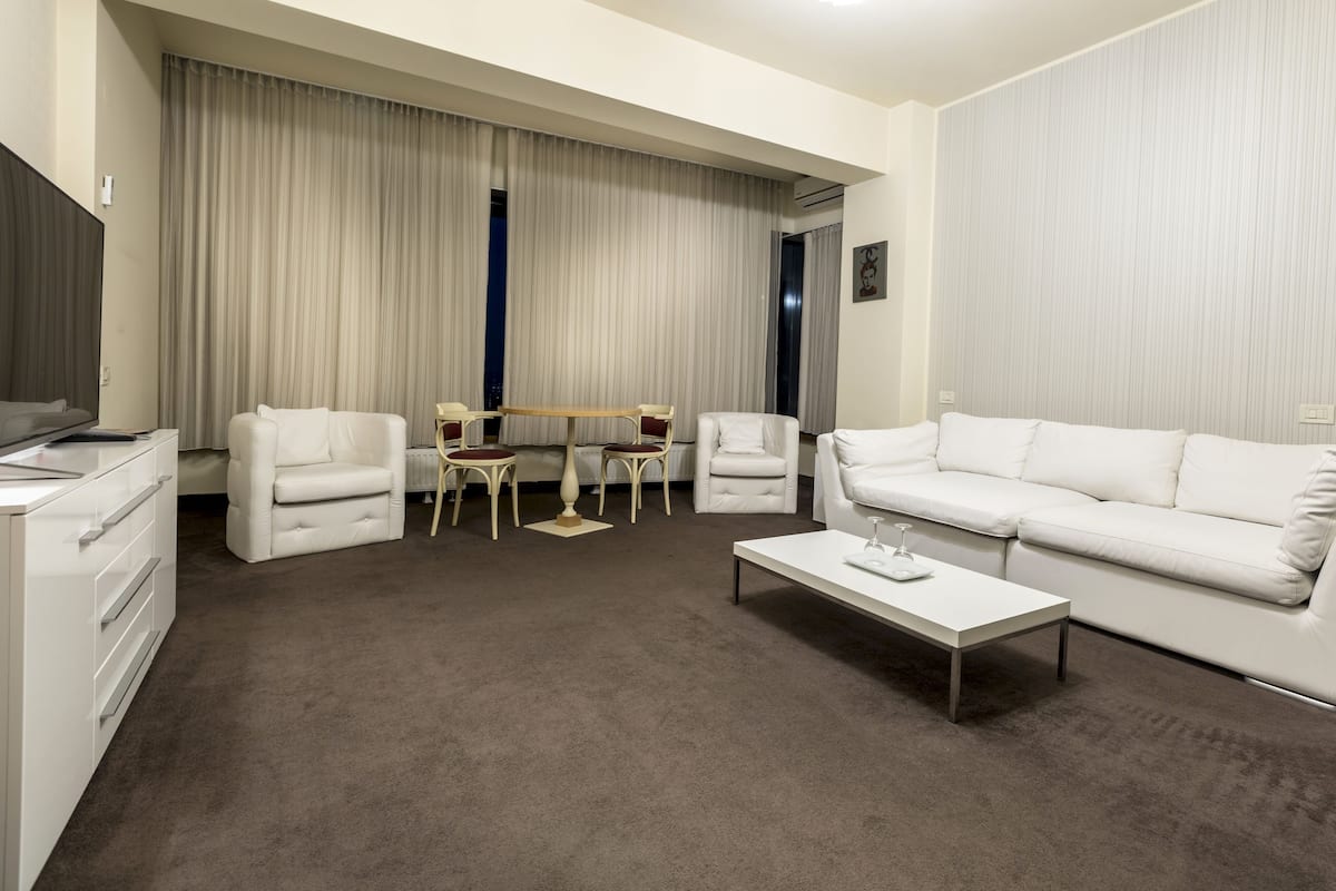 A modern hotel room with a white sofa, a coffee table, a dining table with chairs, and a large window with curtains.