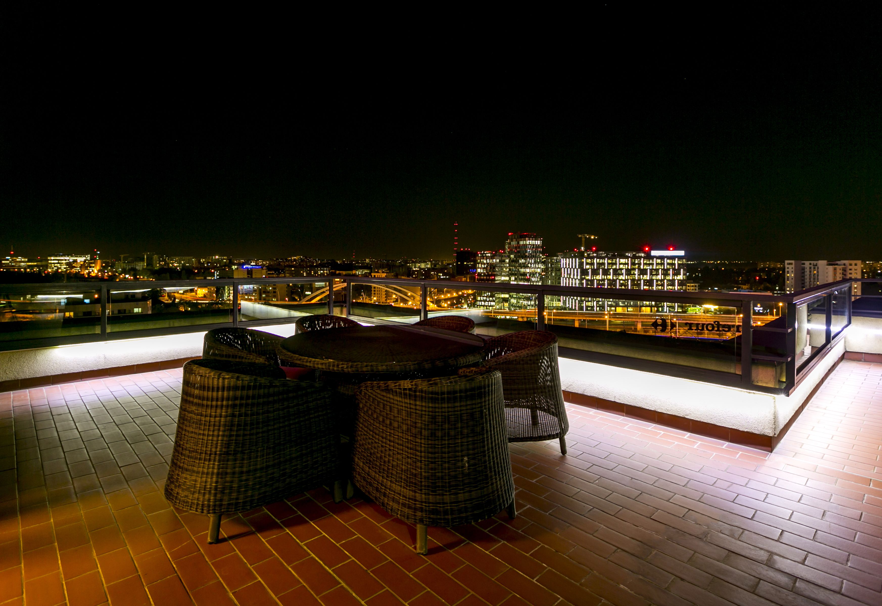 A rooftop terrace with wicker furniture and a city view at night.
