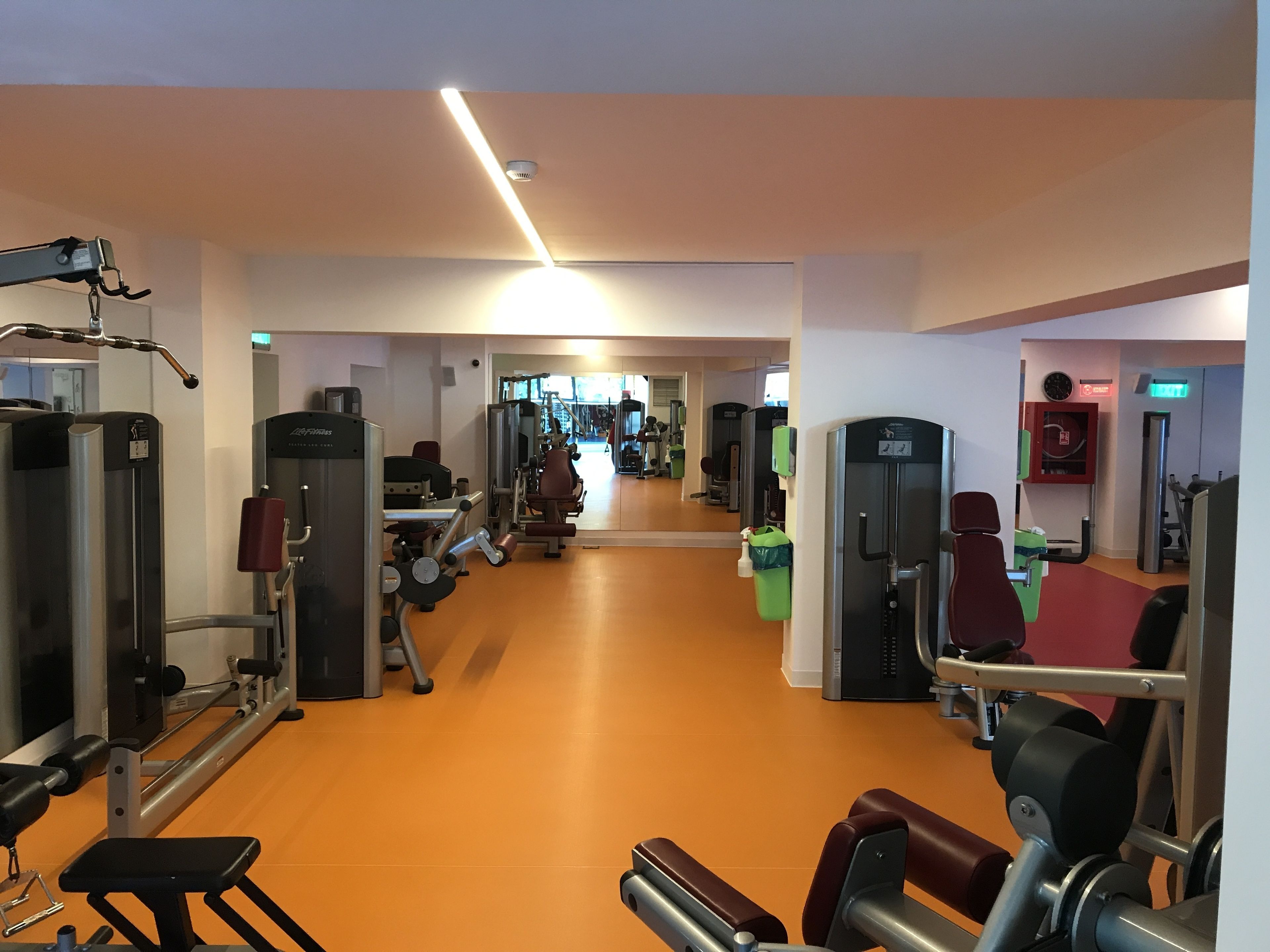 A gym with various exercise machines, a mirrored wall, and a bright overhead light.