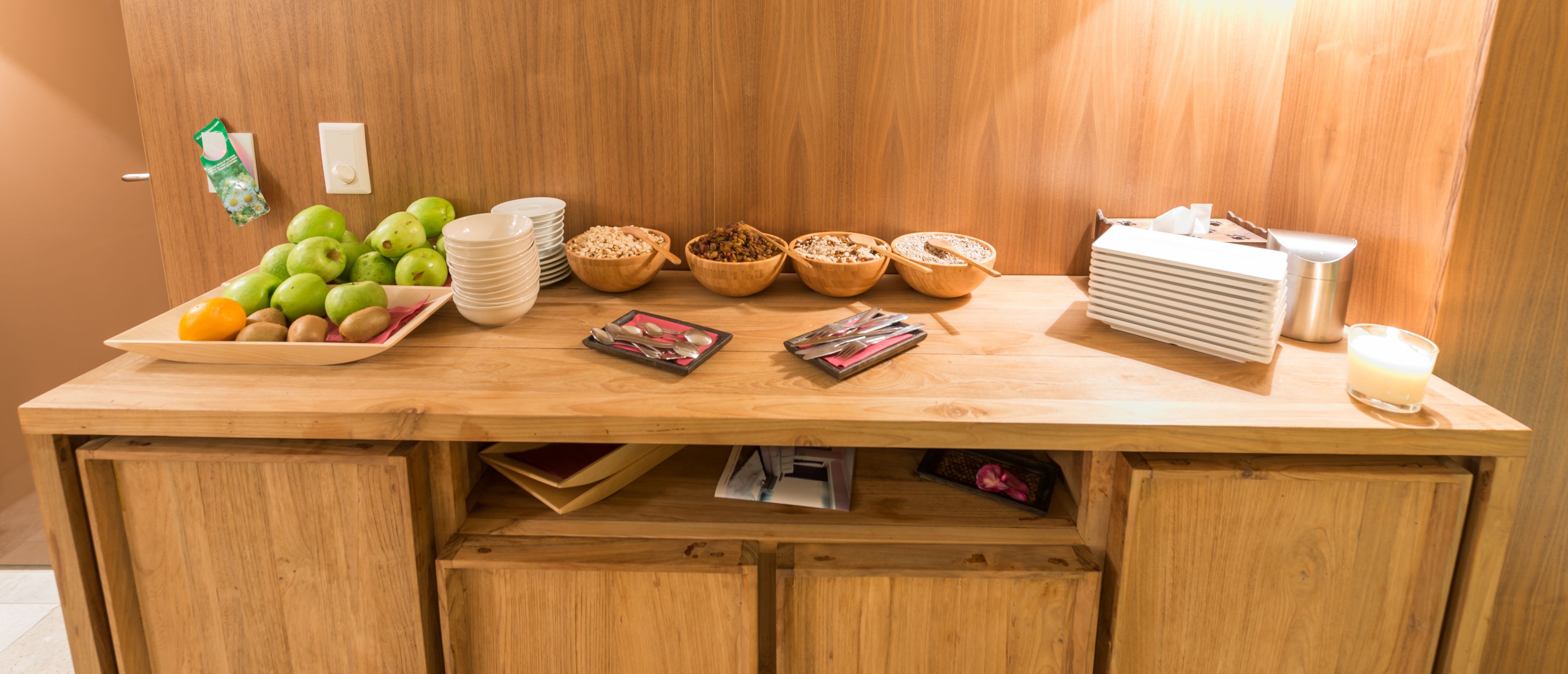 A wooden buffet table with various food items, including fruits, bowls of cereal, and stacked plates.