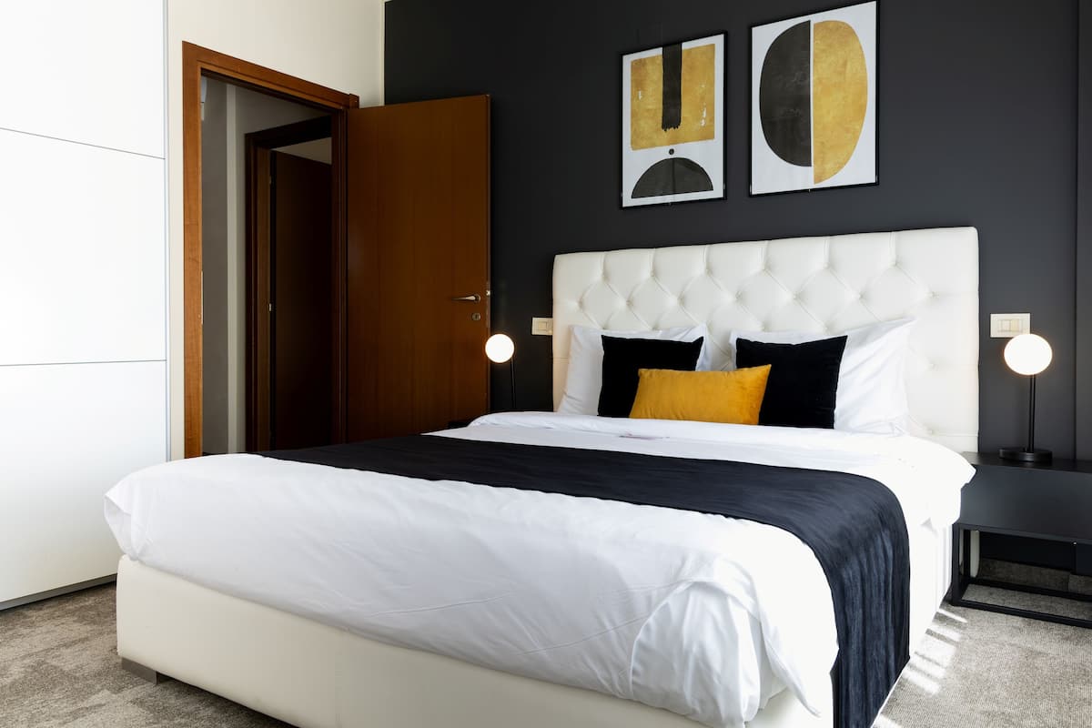 A modern bedroom with a white tufted headboard, a large bed with white and black bedding, a yellow pillow, and two wall-mounted abstract art pieces.