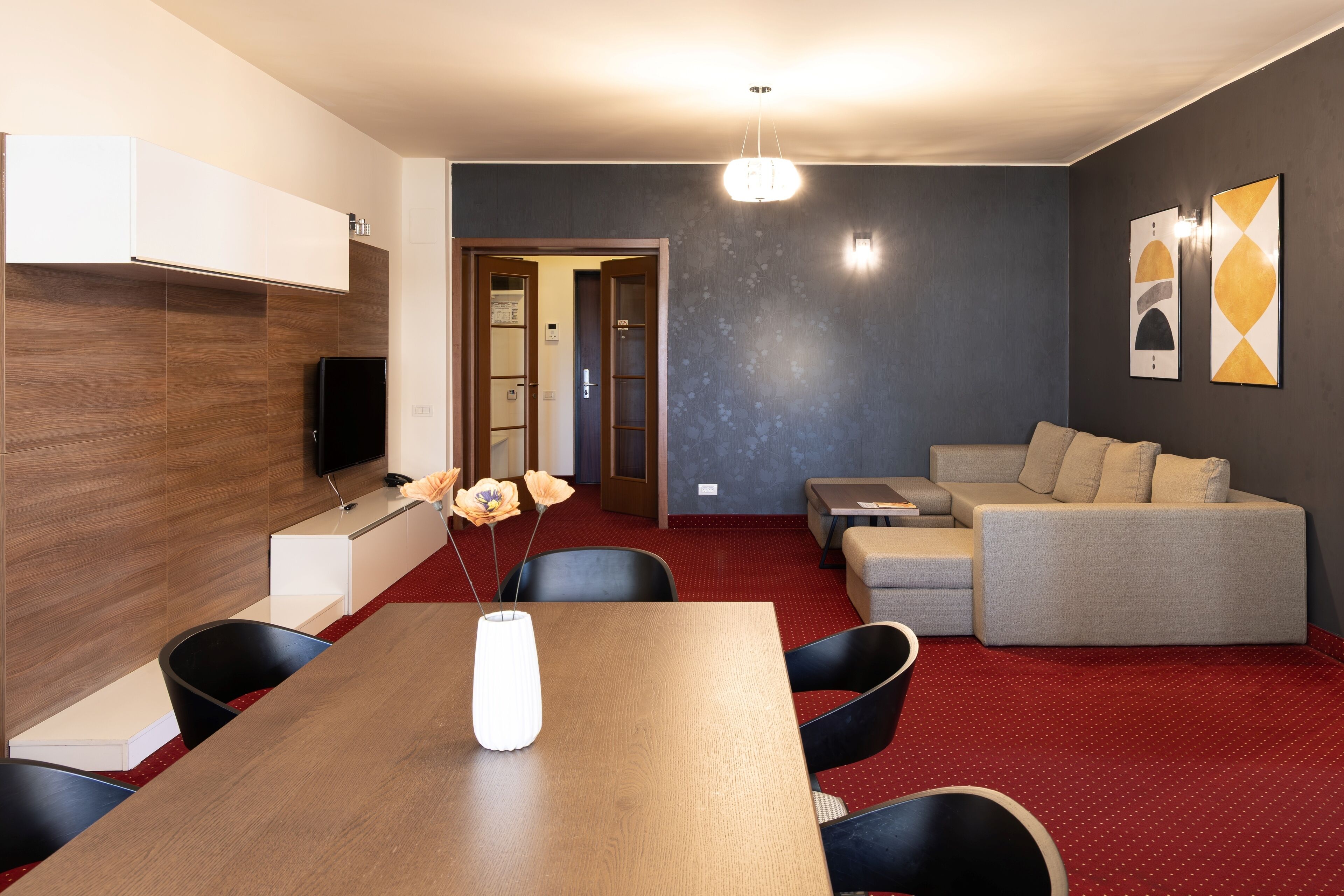 A modern living room with a wooden dining table, black chairs, a grey sofa, and a red carpet.