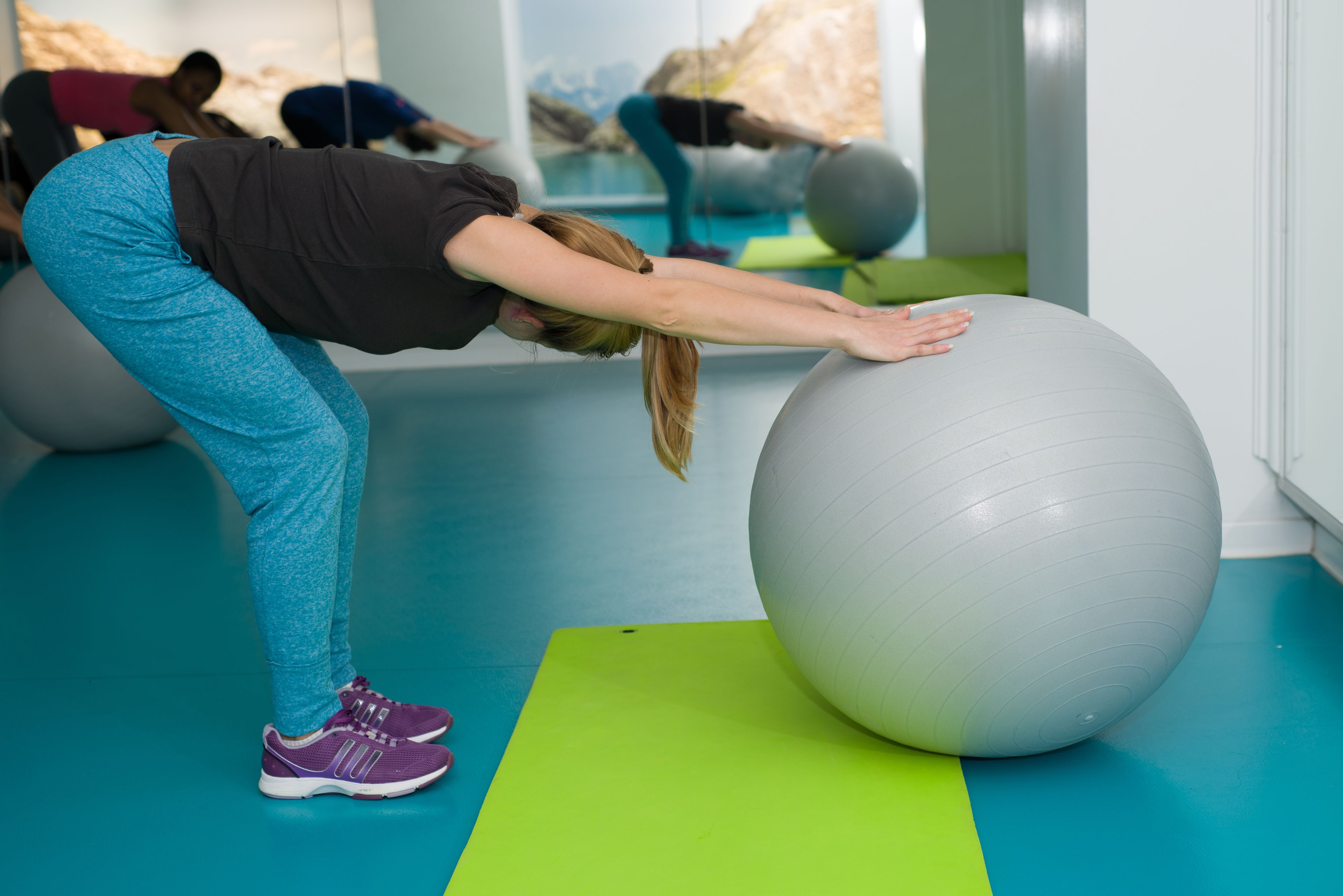 A person performing a stretching exercise on a large exercise ball in a gym with blue flooring and green mats.