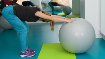 A person performing a stretching exercise on a large exercise ball in a gym with blue flooring and green mats.