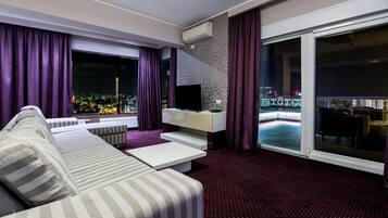 Une chambre d’hôtel moderne dotée d’un grand canapé, d’un téléviseur à écran plat, d’une petite table et d’un balcon offrant une vue sur les lumières de la ville.
