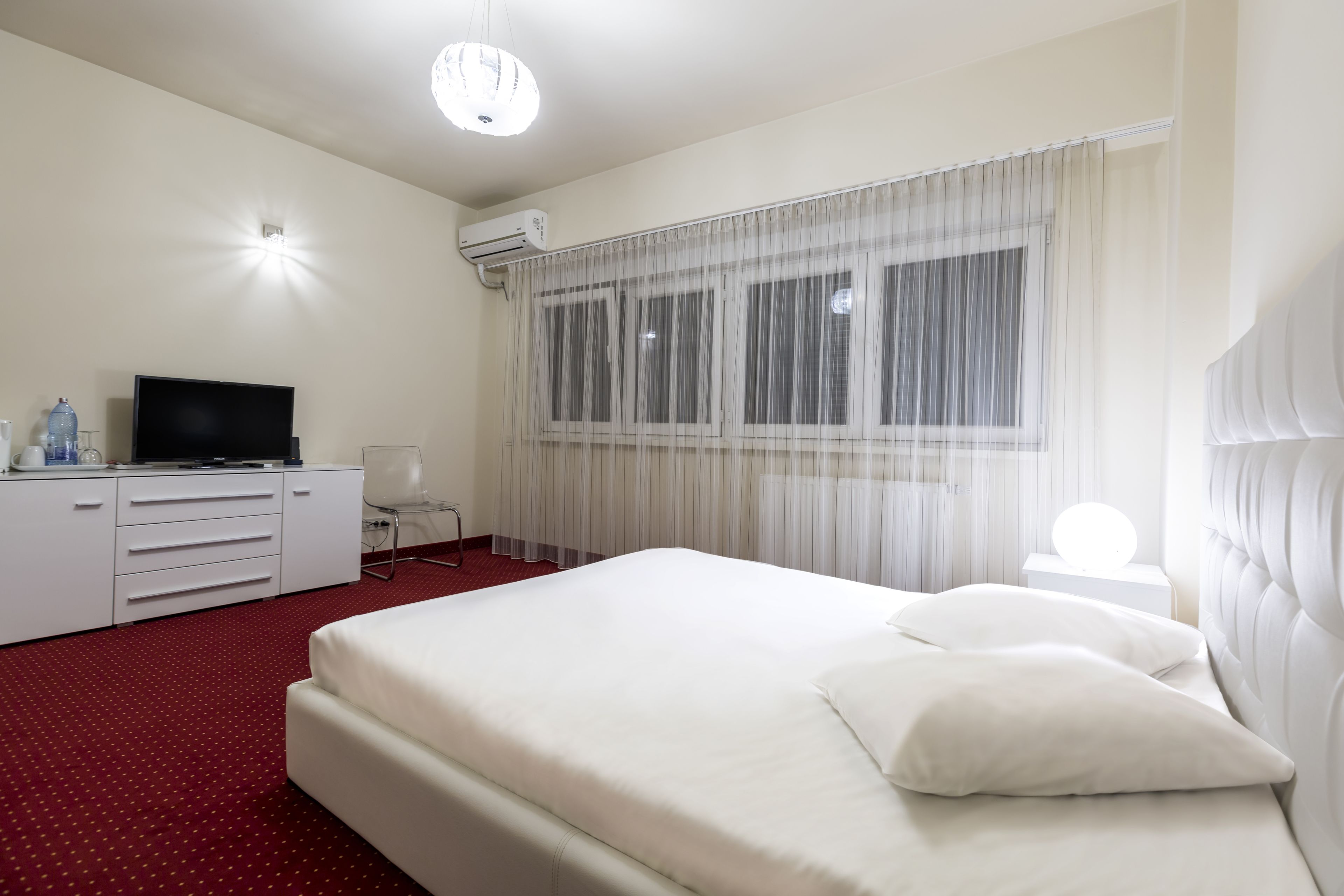 A hotel room with a large bed, a flat-screen TV, a desk with drawers, a water dispenser, and an air conditioning unit.