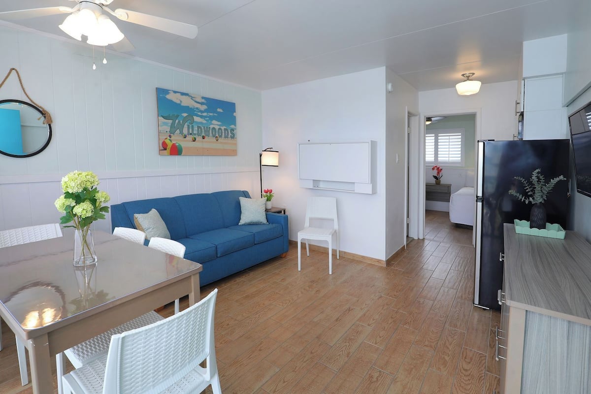 A modern living room with a blue sofa, white chairs, a dining table, and a kitchen area.
