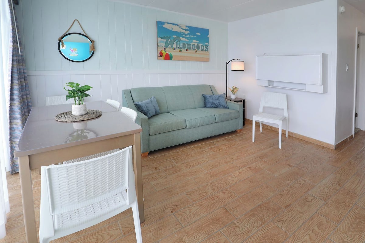 A room with a light green sofa, a white chair, a small table with a plant, a framed picture on the wall, and a whiteboard.